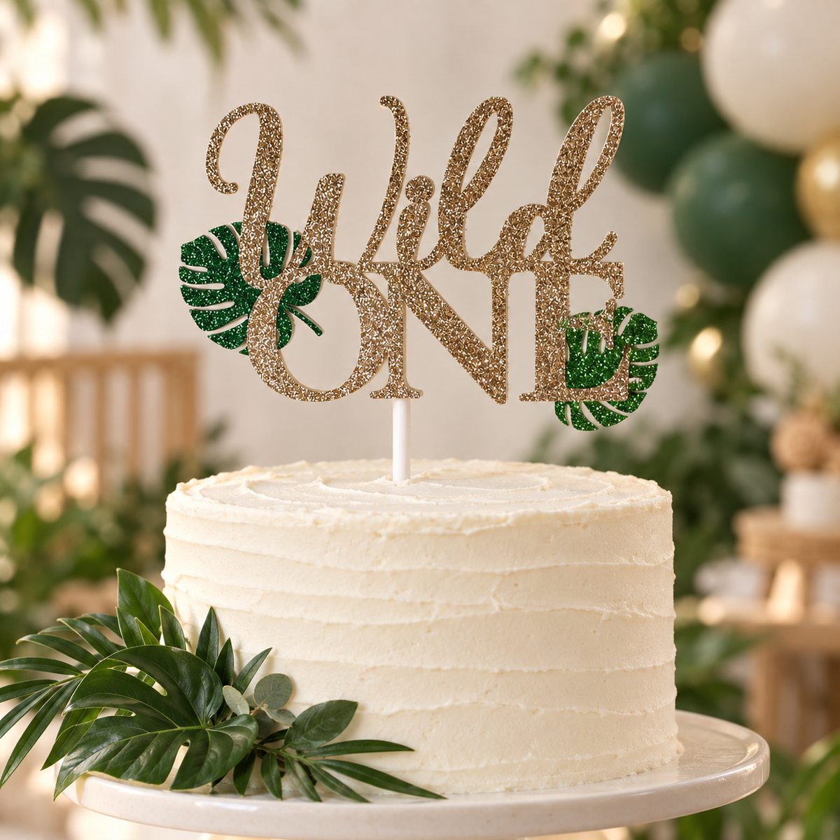Handmade Wild One first birthday cake topper with gold glitter lettering and green tropical leaf accents for a jungle or safari birthday cake.
